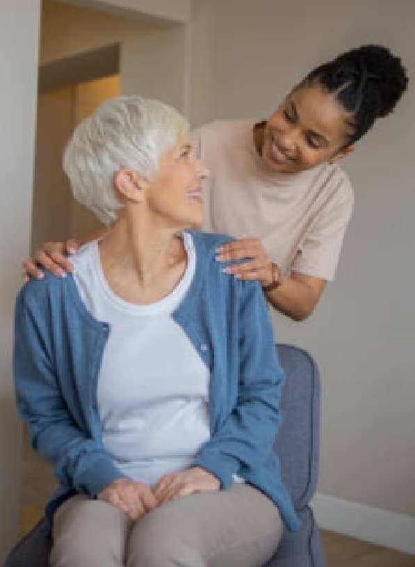 A caregiver standing behind a seated senior woman, hands resting on her shoulders as the two smile at each other at home